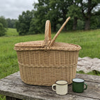 Handwoven wicker picnic basket with one lid lifted on a rustic wooden table outdoors, with two enamel mugs and a blanket on a green meadow