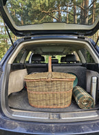 Unpeeled willow picnic basket packed in the open boot of an SUV next to a thermos and a folded tartan blanket for a car trip.