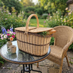 Oval wicker picnic basket for two with lid on a glass garden table beside a rattan chair, flowers and a coffee cup.