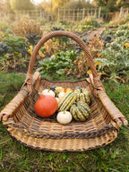 Handwoven willow garden trug with a tall handle filled with pumpkins and squash on the grass beside an autumn vegetable garden