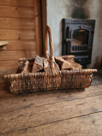 Wicker log carrier with a tall handle filled with chopped firewood on a plank floor beside a cast iron wood stove