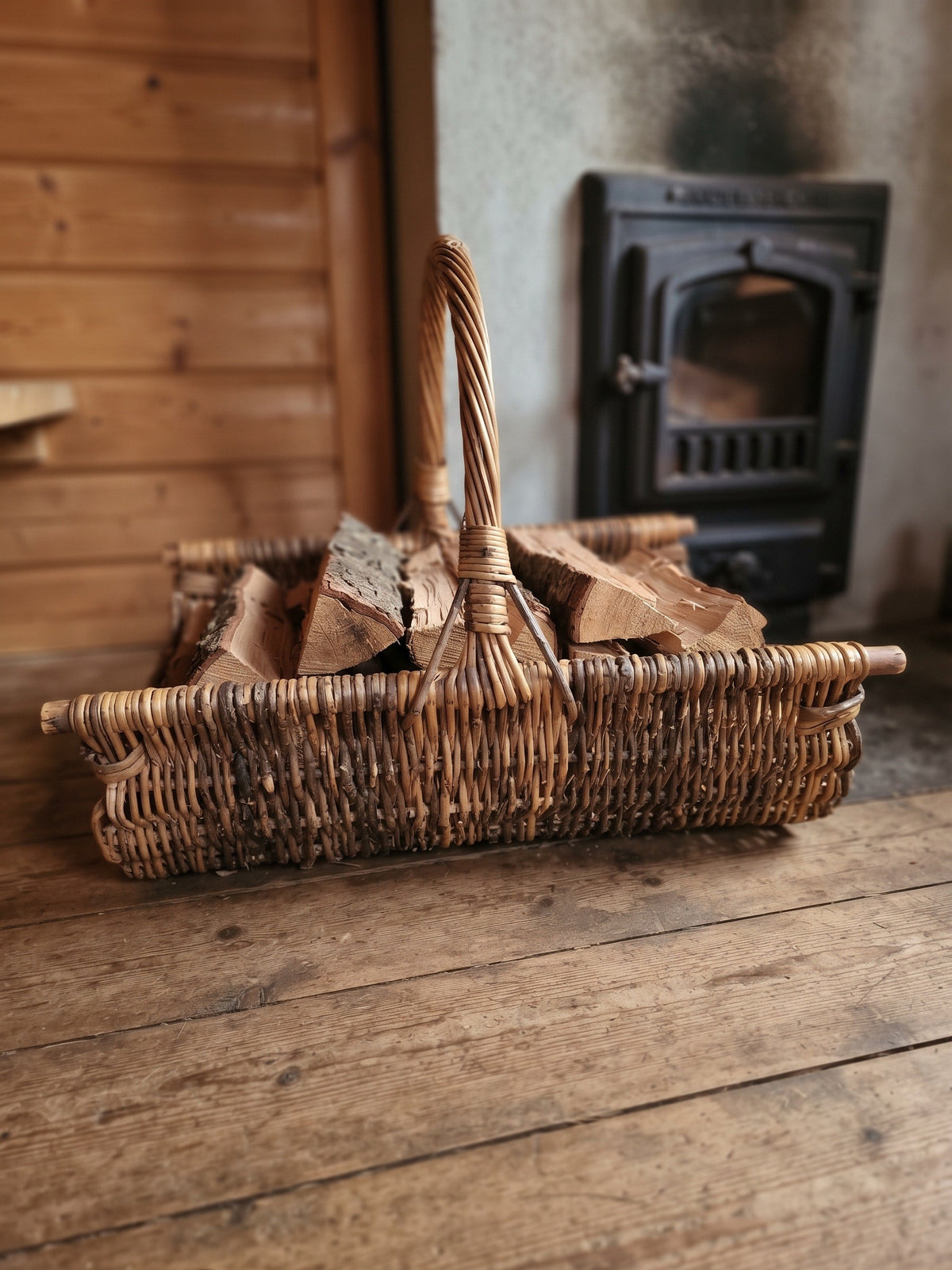 Wicker log carrier with a tall handle filled with chopped firewood on a plank floor beside a cast iron wood stove