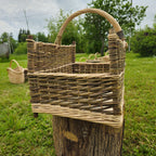Side view of the rectangular willow log basket with corner posts and a high woven handle, standing on a tree stump in the garden