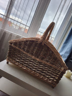 Willow log basket with curved sloping ends and a woven handle, standing on small feet on a white surface by a bright window