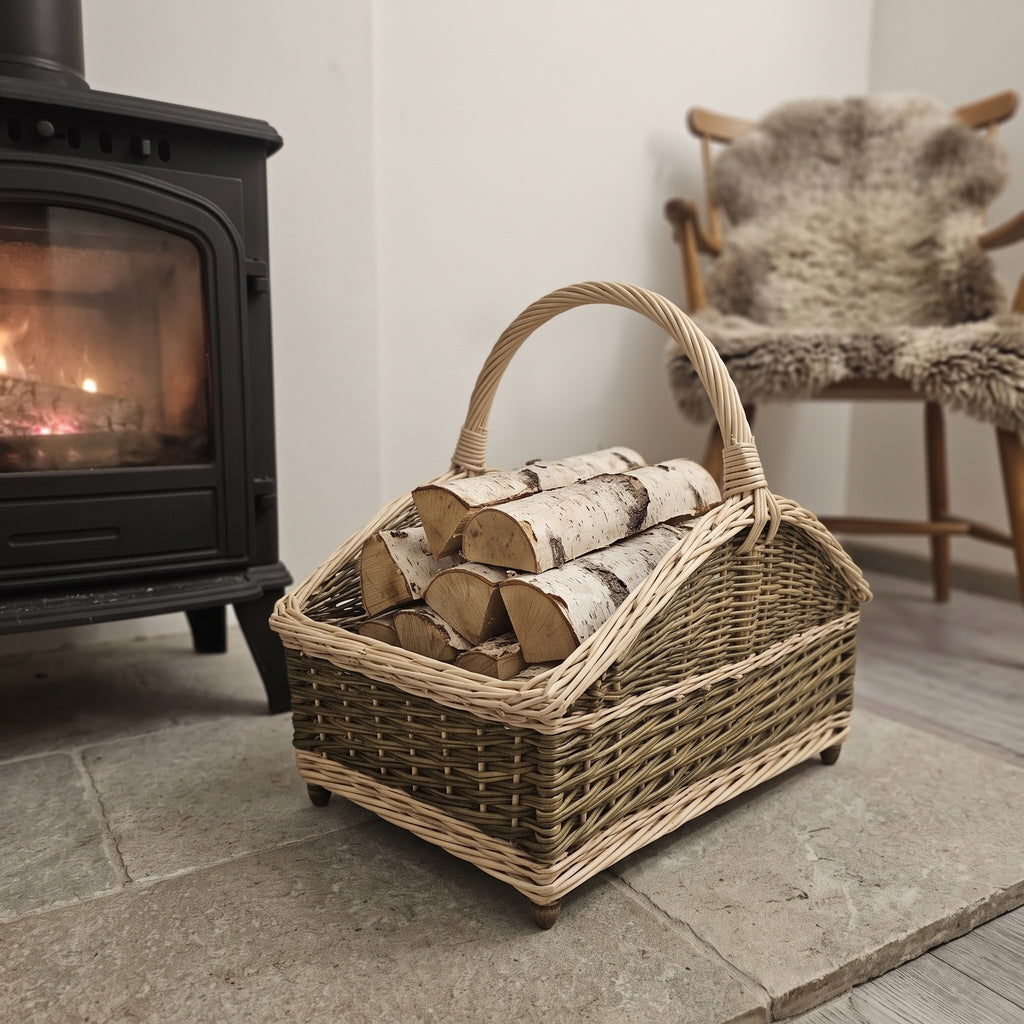 Wicker firewood basket with curved ends and a woven handle, holding birch logs next to a wood-burning stove and a chair with a sheepskin