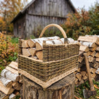 Rectangular willow log basket with corner posts and a handle, packed with birch firewood on a tree stump beside a woodpile and an old barn