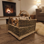 Large handwoven willow log basket with corner posts, stacked with birch firewood on a wooden floor in a living room with a lit fireplace