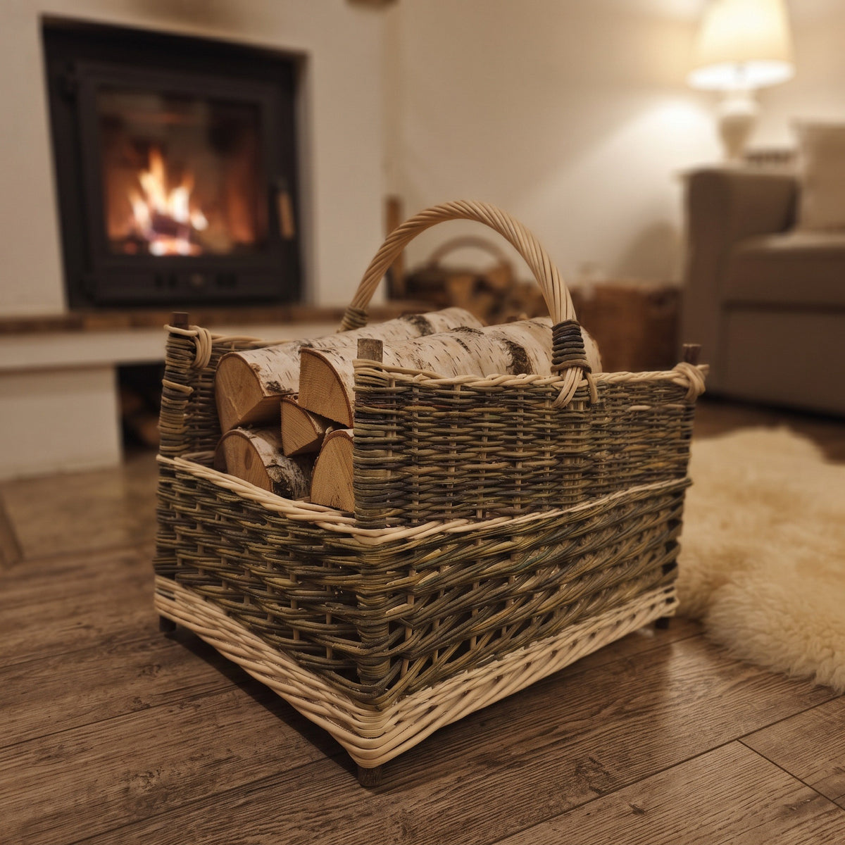 Large handwoven willow log basket with corner posts, stacked with birch firewood on a wooden floor in a living room with a lit fireplace