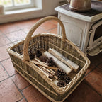 Handwoven willow fireplace basket with curved ends and an arched handle, holding pinecones, kindling sticks and rolled paper beside a stove