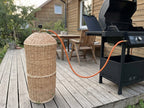 Wicker gas bottle sleeve woven from willow standing on a wooden terrace with the hose running to a BBQ grill outside a timber house