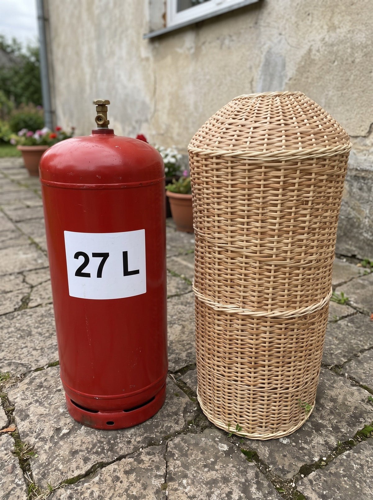 60 cm wicker gas bottle cover beside a red 27 L gas cylinder on a stone patio, showing the smaller size of the willow sleeve