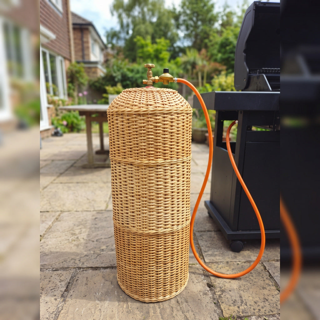 Wicker propane tank sleeve with an orange gas hose connected at the top, standing on stone paving next to a BBQ in a sunny garden
