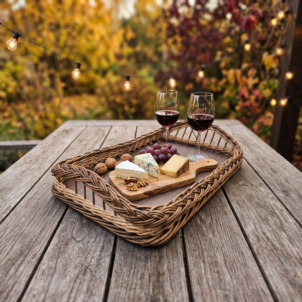 Large wicker breakfast tray with wooden base and braided rim, set with red wine, cheese, grapes and walnuts on a garden table