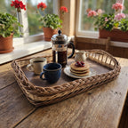 Handwoven willow breakfast tray with coffee, a French press, milk jug and pancakes on a wooden table by a window with geraniums