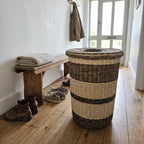 Tall wicker laundry hamper with lid standing in a bright hallway beside a wooden bench with folded towels and boots