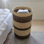 Tall willow laundry basket without its lid holding folded laundry on a rug beside a bed in a bright bedroom