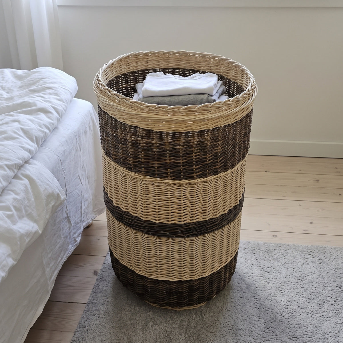 Tall willow laundry basket without its lid holding folded laundry on a rug beside a bed in a bright bedroom