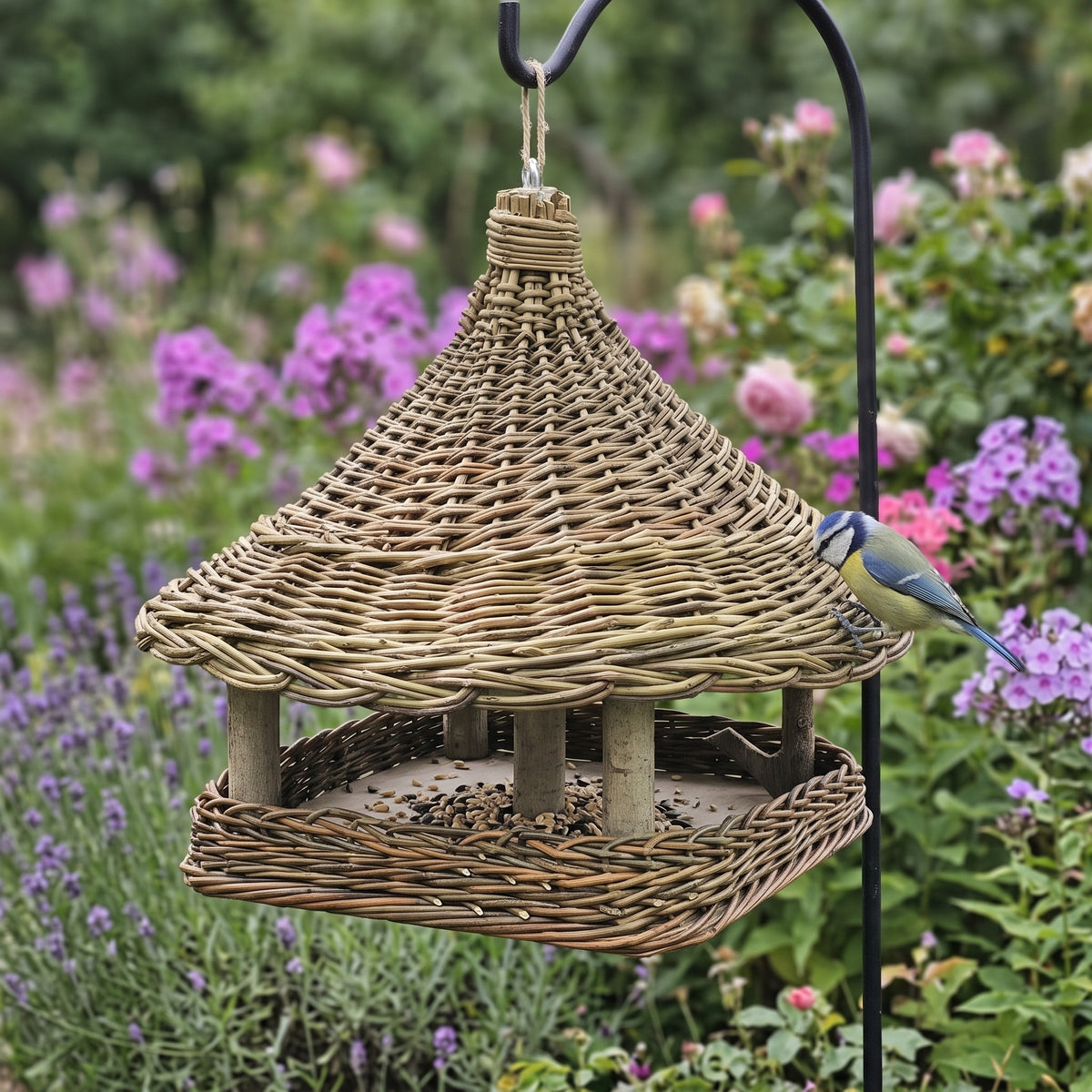 Hanging bird feeder with a cone-shaped woven roof and square seed tray, handwoven from willow, hanging from a shepherd's hook in a flower garden with a blue tit.