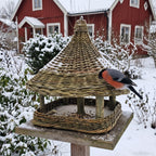 Wicker bird feeder with a cone roof mounted on a wooden post in a snowy garden, a bullfinch perched on the edge of the seed tray.