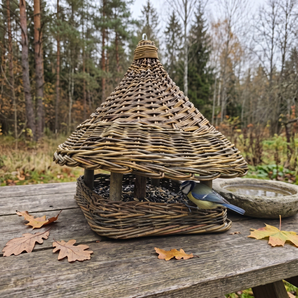 Handwoven willow bird feeder with a woven roof and square tray standing on a garden table among autumn leaves, a blue tit feeding on seed.