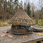 Handwoven willow bird feeder with a woven roof and square tray standing on a garden table among autumn leaves, a blue tit feeding on seed.