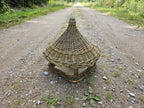Handwoven bird feeder seen down a gravel country track, the woven roof catching the light.