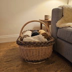 Large wicker blanket basket with handle holding folded throws on a wooden floor next to a sofa in a living room