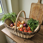 Large oval wicker vegetable basket with handle full of carrots, onions, tomatoes and herbs on a kitchen counter