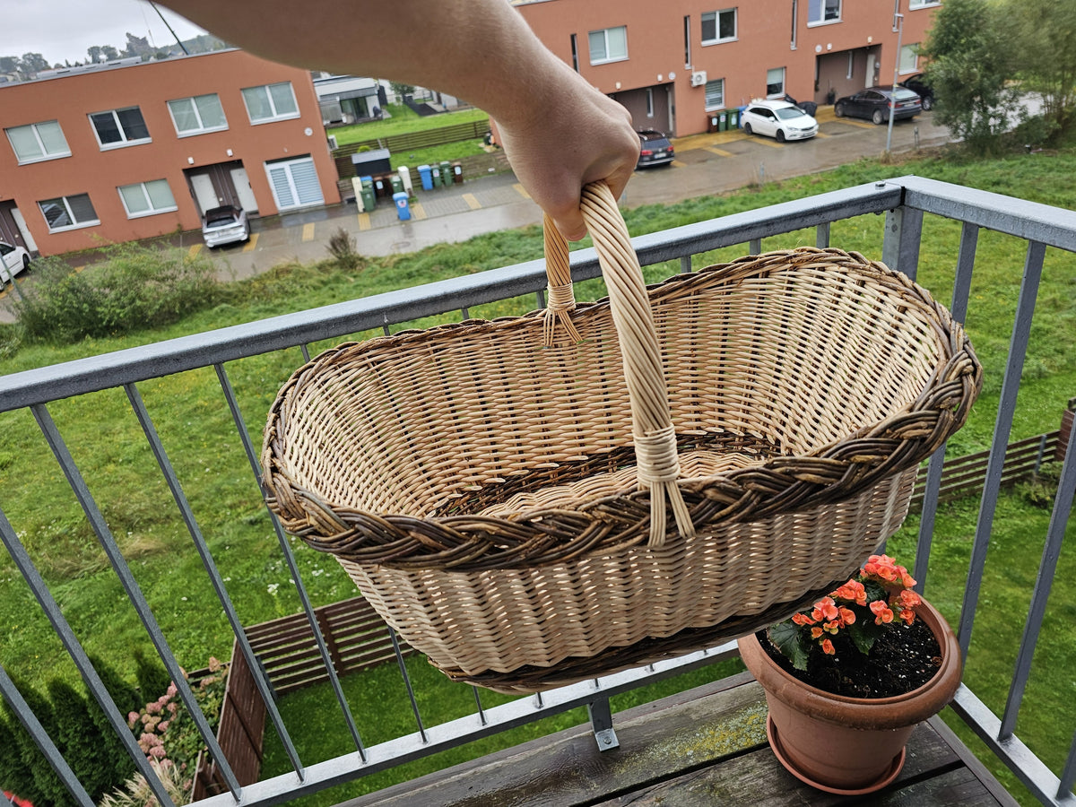 Large oval wicker shopping basket with a tall handle held up by hand on a balcony, showing its roomy oval shape