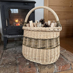 Tall wicker storage basket with handle filled with kindling and pinecones on a brick hearth beside a wood-burning stove