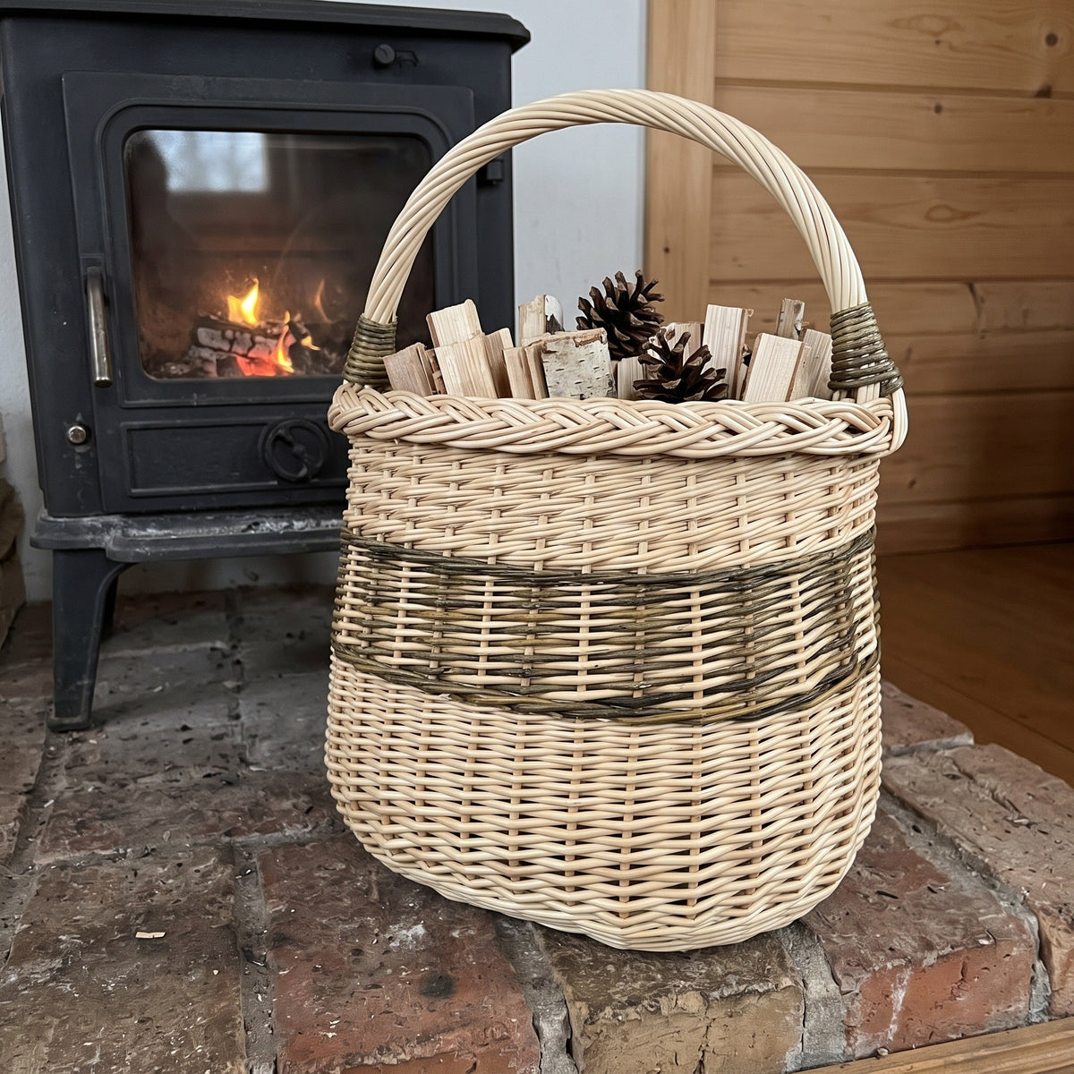 Tall wicker storage basket with handle filled with kindling and pinecones on a brick hearth beside a wood-burning stove