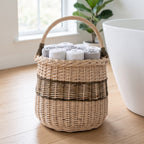 Tall handwoven willow storage basket with handle holding rolled white towels on a wooden floor next to a bathtub