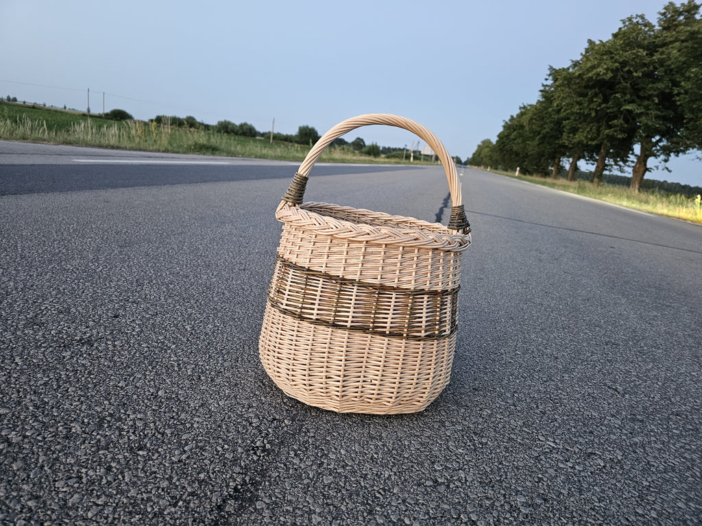 Tall wicker basket with an arched handle photographed on an open asphalt road at dusk, showing its rounded handwoven willow body and ribbed bands.