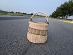 Tall wicker basket with an arched handle photographed on an open asphalt road at dusk, showing its rounded handwoven willow body and ribbed bands.