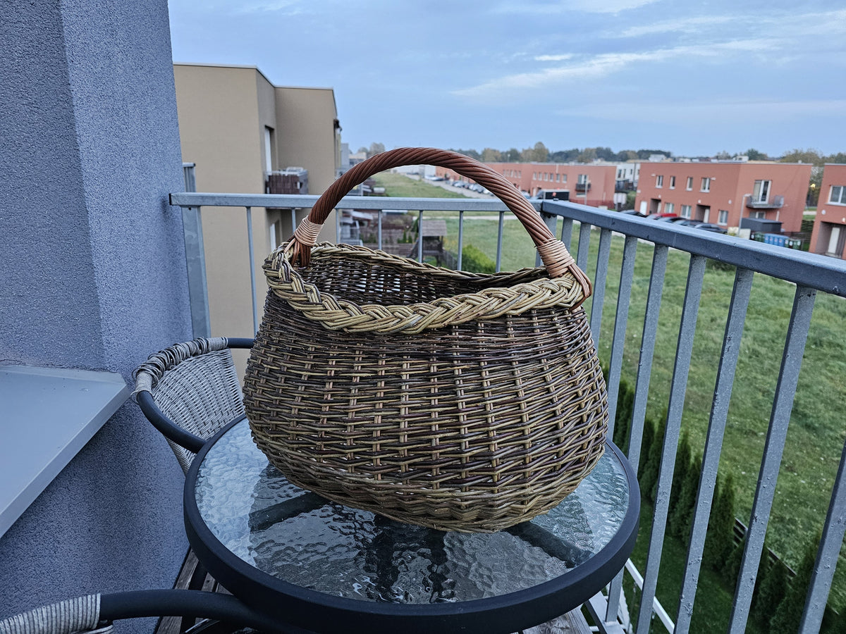 Handwoven willow foraging basket with a braided rim on a round glass table on a city balcony, apartment buildings in the background.