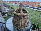 Oval willow foraging basket with a tall wrapped handle on a glass balcony table, showing its deep rounded body and braided rim.