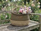 Deep wicker basket holding a bouquet of sweet peas and roses on a weathered garden bench in a flowering cottage garden.