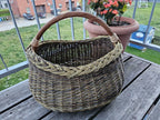 Handwoven oval foraging basket with braided rim on a wooden balcony table beside a pot of flowering begonias.