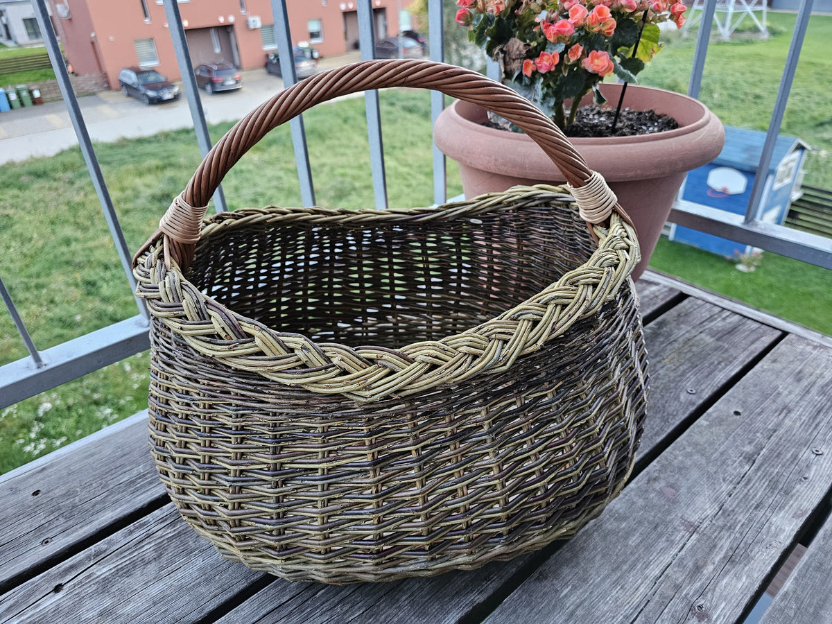 Handwoven oval foraging basket with braided rim on a wooden balcony table beside a pot of flowering begonias.