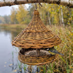 Handwoven willow bird feeder with a cone roof hanging from a birch branch by a lake, a great tit perched on the round seed tray among autumn reeds.