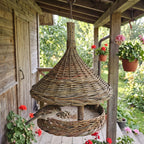 Willow bird feeder with a tall woven top hanging from a wooden porch beam, a sparrow eating seed from the tray beside pots of red geraniums.