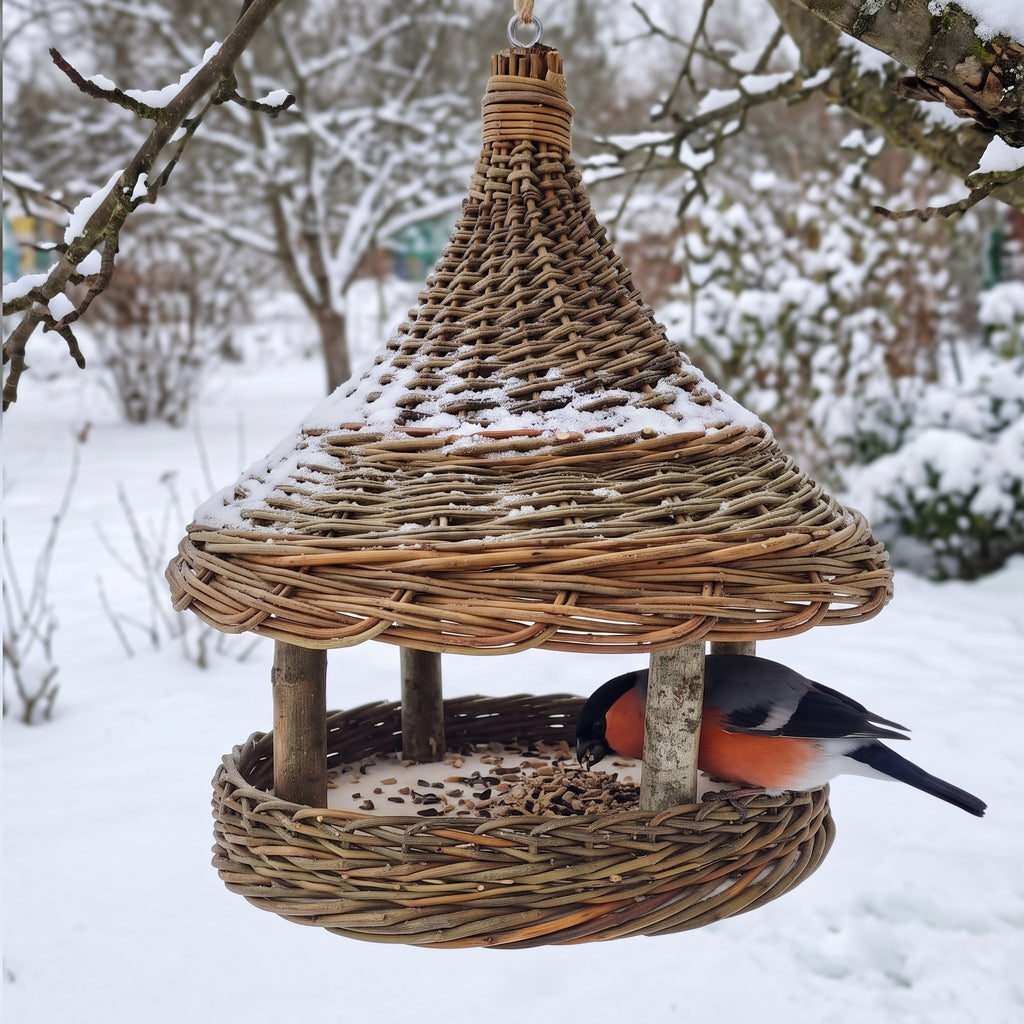 Hanging willow seed tray feeder in a snowy garden, snow resting on the cone roof while a bullfinch eats sunflower seeds from the round tray.