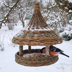 Hanging willow seed tray feeder in a snowy garden, snow resting on the cone roof while a bullfinch eats sunflower seeds from the round tray.
