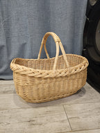 Oval handwoven willow foraging basket with twin arched handles joined at the top, standing on a wooden floor against a grey curtain