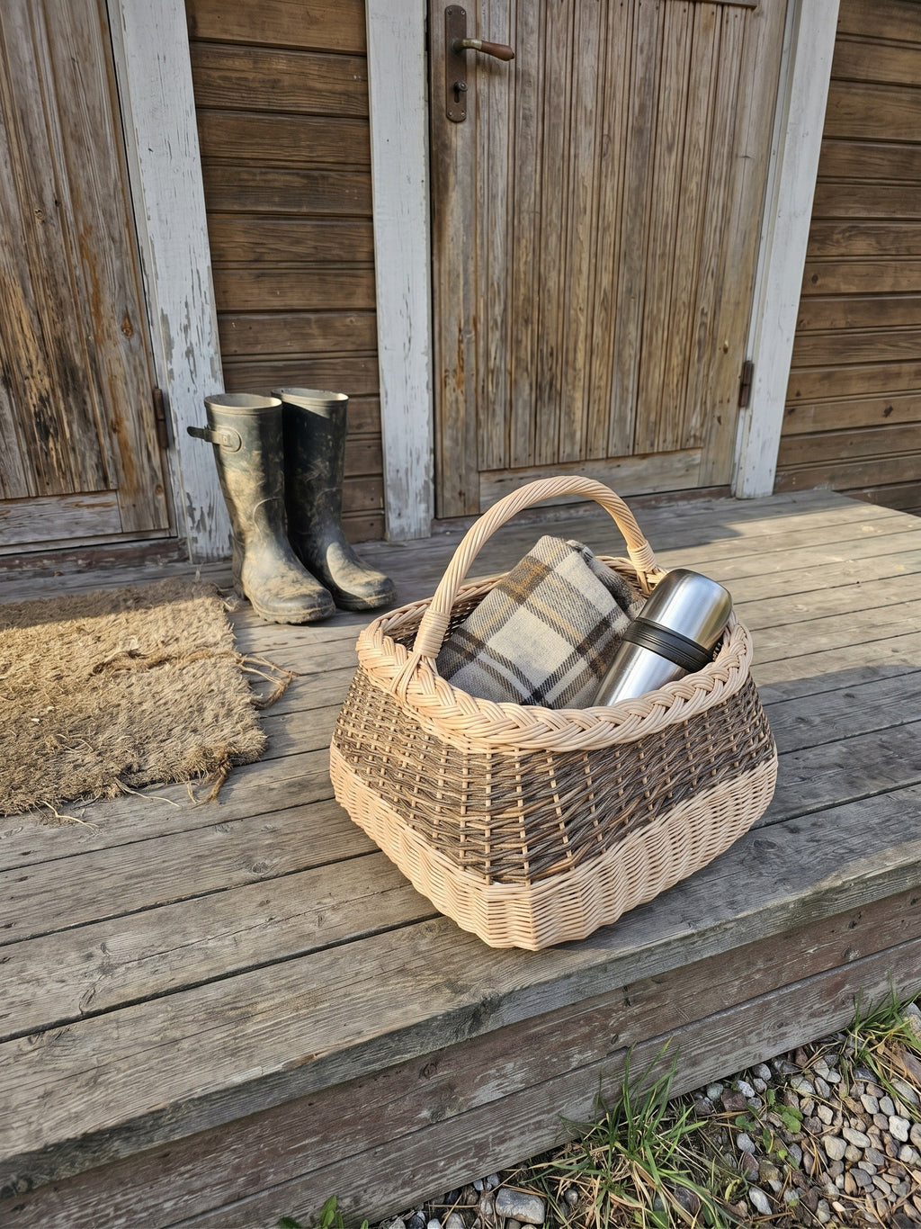 Large square gathering basket with handle, handwoven from willow with a braided rim, holding a wool blanket and thermos on a wooden porch beside rubber boots.