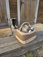 Large square gathering basket with handle, handwoven from willow with a braided rim, holding a wool blanket and thermos on a wooden porch beside rubber boots.