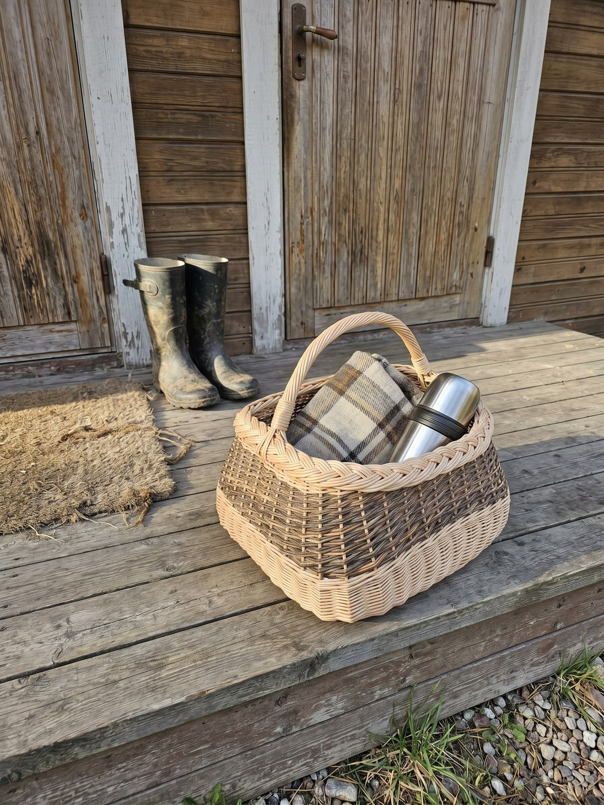 Large square gathering basket with handle, handwoven from willow with a braided rim, holding a wool blanket and thermos on a wooden porch beside rubber boots.