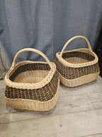 Two handmade willow gathering baskets in different sizes shown side by side indoors, both with braided rims, chevron weave bands and tall arched handles.