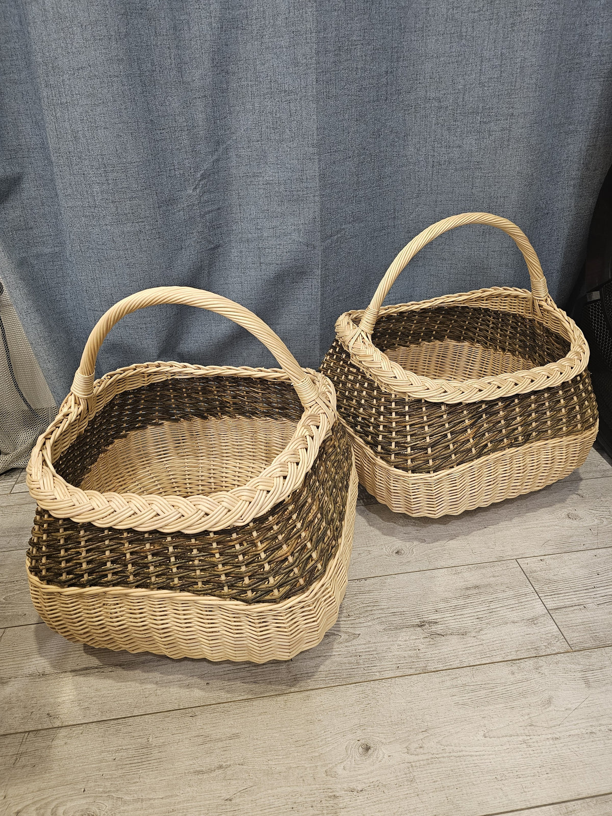 Two handmade willow gathering baskets in different sizes shown side by side indoors, both with braided rims, chevron weave bands and tall arched handles.