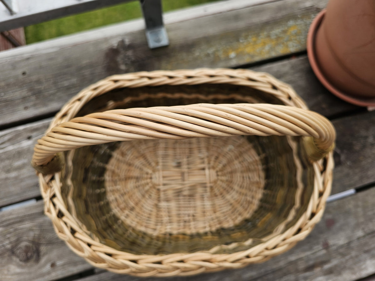 Top-down view of an oval willow market basket on a wooden table, showing the handle across the braided rim and the woven oval base inside.